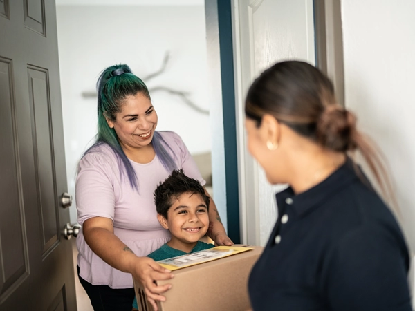 A Hispanic mother and her son receive a package delivered by another woman at the door.