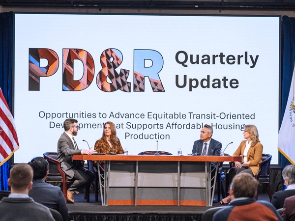 A stage with four panelists sitting at a table. A large screen behind them reads "PD&R Quarterly Update: Opportunities to Advance Equitable Transit-Oriented Development that Supports Affordable Housing Production."